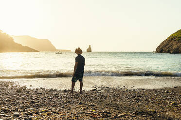 Rückenansicht eines Mannes, der am Strand steht und bei Sonnenuntergang aufs Meer schaut, Cala Benirras, Ibiza, Spanien - KIJF02996