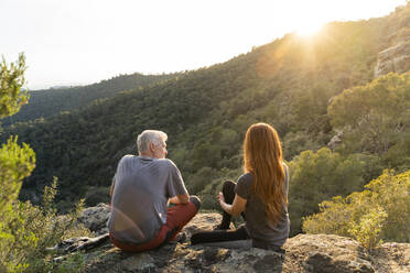 Rückenansicht von Vater und erwachsener Tochter, die auf einem Felsen in den Bergen sitzen und den Sonnenuntergang genießen - AFVF06206