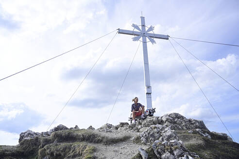 Niedriger Blickwinkel auf eine Frau, die am Gipfelkreuz des Kramerspitzgipfels sitzt, gegen den Himmel - ECPF00905