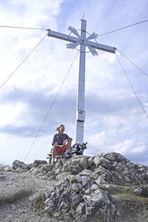 Low angle view of woman sitting on bench by summit cross at Kramerspitz mountain peak against sky - ECPF00906