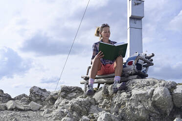 Frau in voller Länge beim Lesen eines Buches, auf einer Bank sitzend, über Felsen am Kramerspitzberg gegen den Himmel - ECPF00907