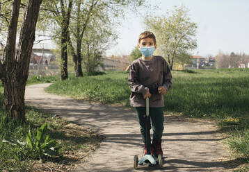 Porträt eines Jungen mit Schutzmaske und Kick-Scooter in einem Park - MOMF00863