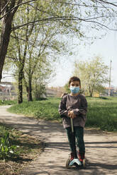 Portrait of boy wearing protective mask with kick scooter in a park - MOMF00864