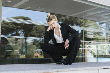 Porträt einer jungen Geschäftsfrau im schwarzen Hosenanzug, die vor einem Bürogebäude hockt - TCEF00576