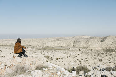Frau in voller Länge auf einem Felsen sitzend mit Blick auf die Wüstenlandschaft und den klaren blauen Himmel - XLGF00125