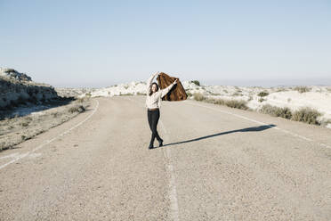 Lächelnde Frau in voller Länge, die ihre Jacke hält, während sie auf der Straße in der Wüste gegen den klaren Himmel läuft - XLGF00126