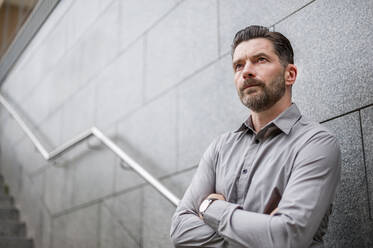 Low angle view of thoughtful businessman standing with arms crossed against wall on staircase in city - DIGF10265