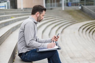 Seitenansicht eines gut aussehenden Geschäftsmannes, der sein Smartphone benutzt, während er mit einer Akte auf den Stufen eines Amphitheaters sitzt - DIGF10267