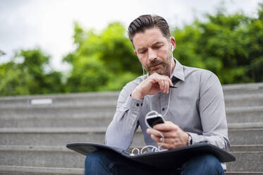 Selbstbewusster Geschäftsmann, der sein Smartphone benutzt, während er mit einer Akte im Amphitheater sitzt - DIGF10268