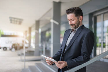 Smiling bearded entrepreneur looking at digital tablet while standing outside office building - DIGF10273