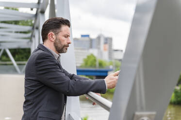 Seitenansicht eines gutaussehenden, bärtigen Unternehmers, der mit einem Mobiltelefon auf einer Fußgängerbrücke in der Stadt steht - DIGF10278