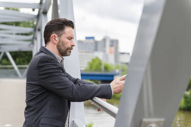 Side view of handsome bearded entrepreneur standing on footbridge with mobile phone in city - DIGF10278