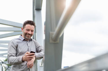 Smiling handsome businessman using smart phone while standing on footbridge in city - DIGF10280