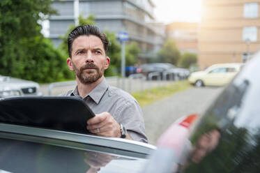 Nachdenklicher Geschäftsmann schaut weg, während er eine Akte auf dem Autodach eines Parkplatzes in der Stadt hält - DIGF10282