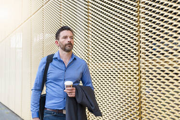 Handsome businessman walking with coffee by wall in city - DIGF10283