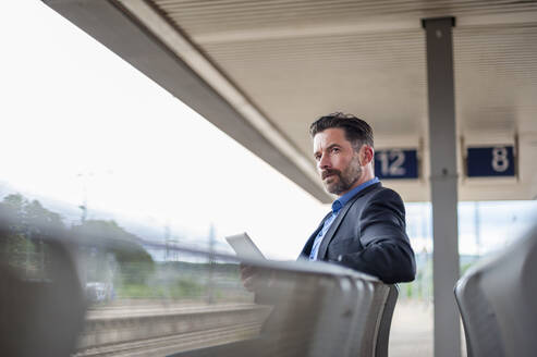 Selbstbewusster Geschäftsmann mit digitalem Tablet sitzend und wegschauend am Bahnhof - DIGF10290