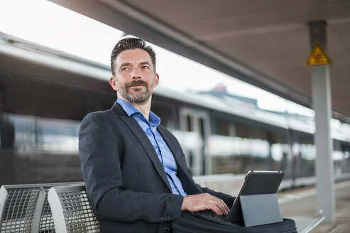 Selbstbewusster Geschäftsmann, der wegschaut, während er mit einem digitalen Tablet am Bahnhof sitzt und wartet - DIGF10293