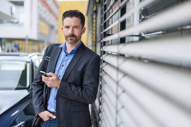 Portrait of confident businessman standing with smart phone in city - DIGF10294