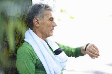 Side view of active senior man looking at smart watch while standing against tree trunk in park - DIGF10311