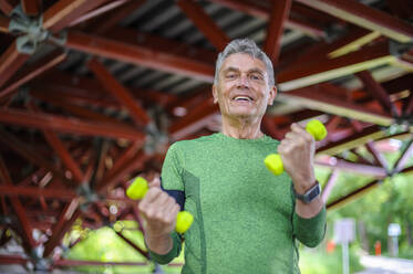 Low angle portrait of smiling senior man exercising with dumbbells while standing at gazebo - DIGF10314