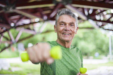 Portrait of smiling senior man exercising with dumbbells while standing at gazebo in park - DIGF10315