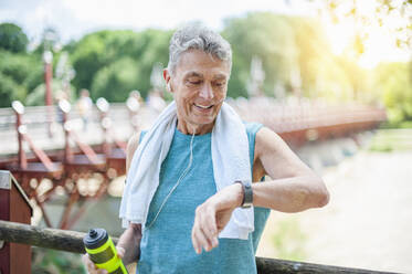 Smiling active senior man looking at smart watch while standing in park - DIGF10319