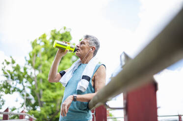 Low angle view of thirsty senior man drinking water from bottle while leaning on railing at park - DIGF10320