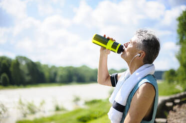 Seitenansicht eines durstigen aktiven älteren Mannes, der am Flussufer an einem sonnigen Tag Wasser aus einer Flasche trinkt - DIGF10323