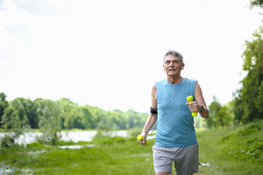 Active senior man jogging with dumbbells at riverbank against clear sky - DIGF10327