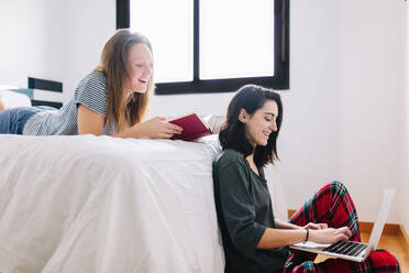 Smiling woman sitting on the floor using laptop while her friend watching from bed - MRRF00003