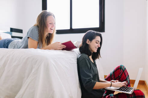 Lächelnde Frau, die auf dem Boden sitzt und einen Laptop benutzt, während ihr Freund vom Bett aus zusieht - MRRF00003