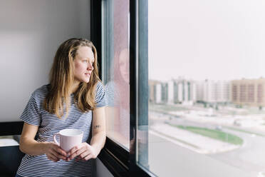 Porträt einer jungen Frau mit Tasse, die aus dem Fenster schaut - MRRF00004