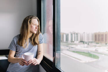 Portrait of young woman with mug looking out of window - MRRF00004