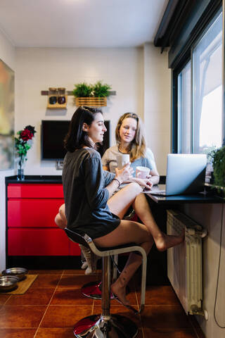 Zwei Frauen sitzen in der Küche und benutzen einen Laptop für einen Videochat, lizenzfreies Stockfoto