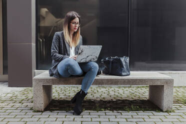 Young freelancer sitting on bench in the city using laptop - MRRF00010