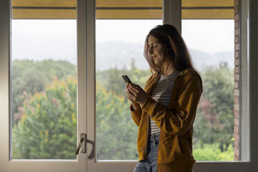 Lächelnde rothaarige Frau steht vor einem Fenster und schaut auf ihr Handy - AFVF06211