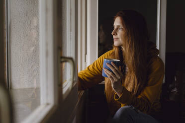 Smiling redheaded woman with coffee mug looking out of open window - AFVF06217