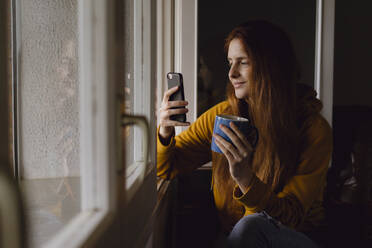 Lächelnde rothaarige Frau mit Kaffeebecher sitzt am offenen Fenster und schaut auf ihr Handy - AFVF06218