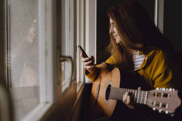 Smiling redheaded woman with guitar sitting at open window looking at cell phone - AFVF06219