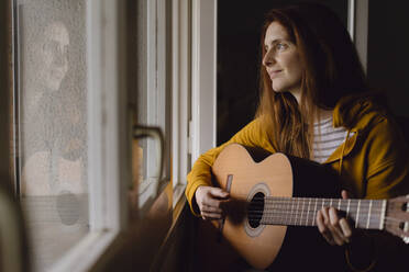 Porträt einer entspannten rothaarigen Frau, die am offenen Fenster Gitarre spielt - AFVF06220