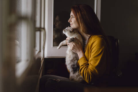 Lächelnde rothaarige Frau sitzt mit ihrer Katze am offenen Fenster zu Hause, lizenzfreies Stockfoto