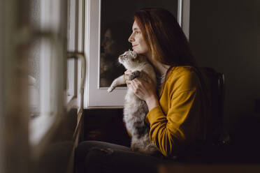 Smiling redheaded woman sitting with her cat at open window at home - AFVF06224