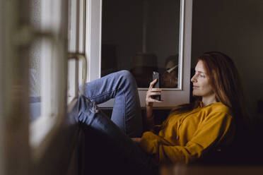 Smiling redheaded woman sitting at open window with feet up looking at cell phone - AFVF06226