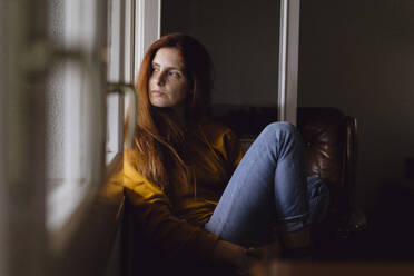 Portrait of redheaded woman sitting at open window looking at home looking at distance - AFVF06227