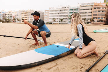 Ein Lehrer zeigt auf eine junge Frau, während er ihr das Paddleboarding am Strand erklärt - MRRF00025