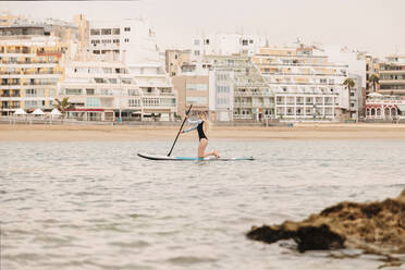 Full length side view of young woman kneeling while practicing paddleboarding on sea - MRRF00031