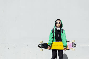 Porträt eines Jungen mit Skateboard und Kopfhörern vor einer weißen Wand stehend - JCMF00697