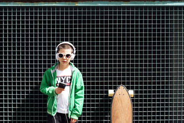 Porträt eines Jungen mit Skateboard, der mit Kopfhörern und Smartphone Musik hört - JCMF00699