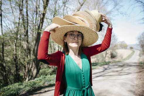 Porträt einer reifen Frau mit Stapel von Strohhüten auf dem Kopf im Freien - KMKF01303