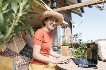 Porträt einer lachenden reifen Frau, die auf einer Terrasse am Laptop arbeitet - KMKF01311